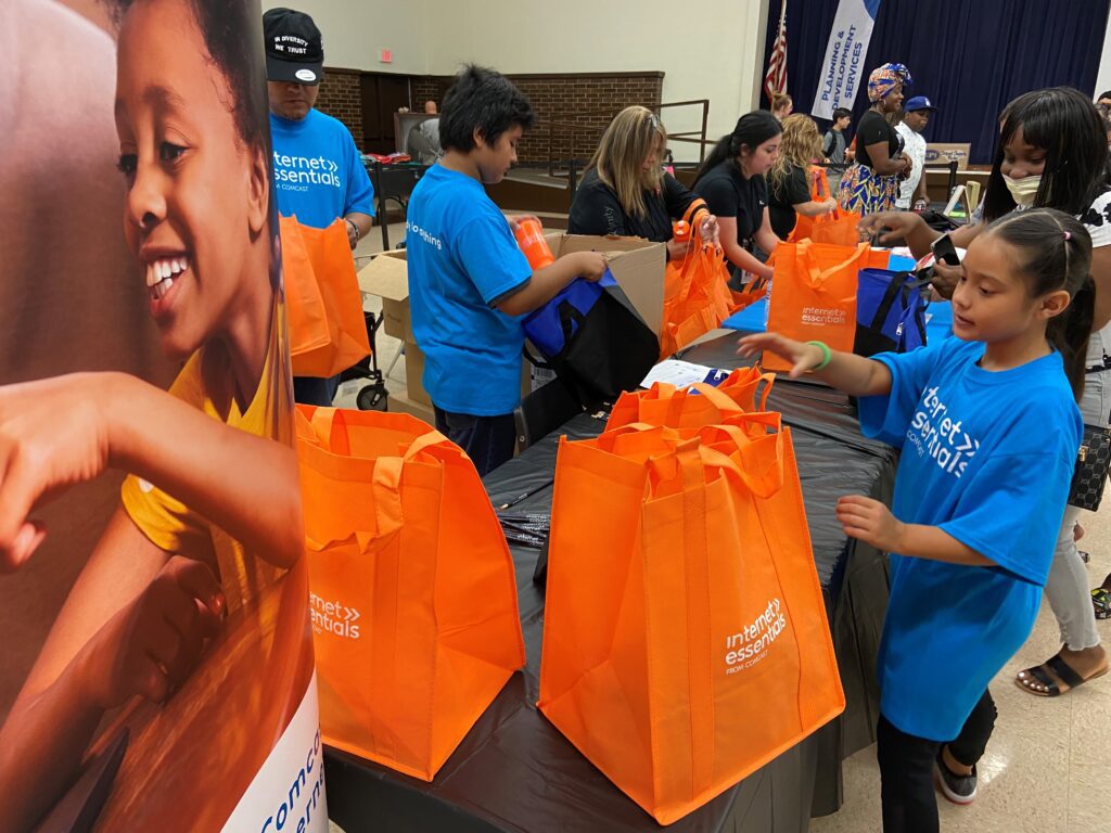 People approaching table containing orange Internet Essentials fabric bags.