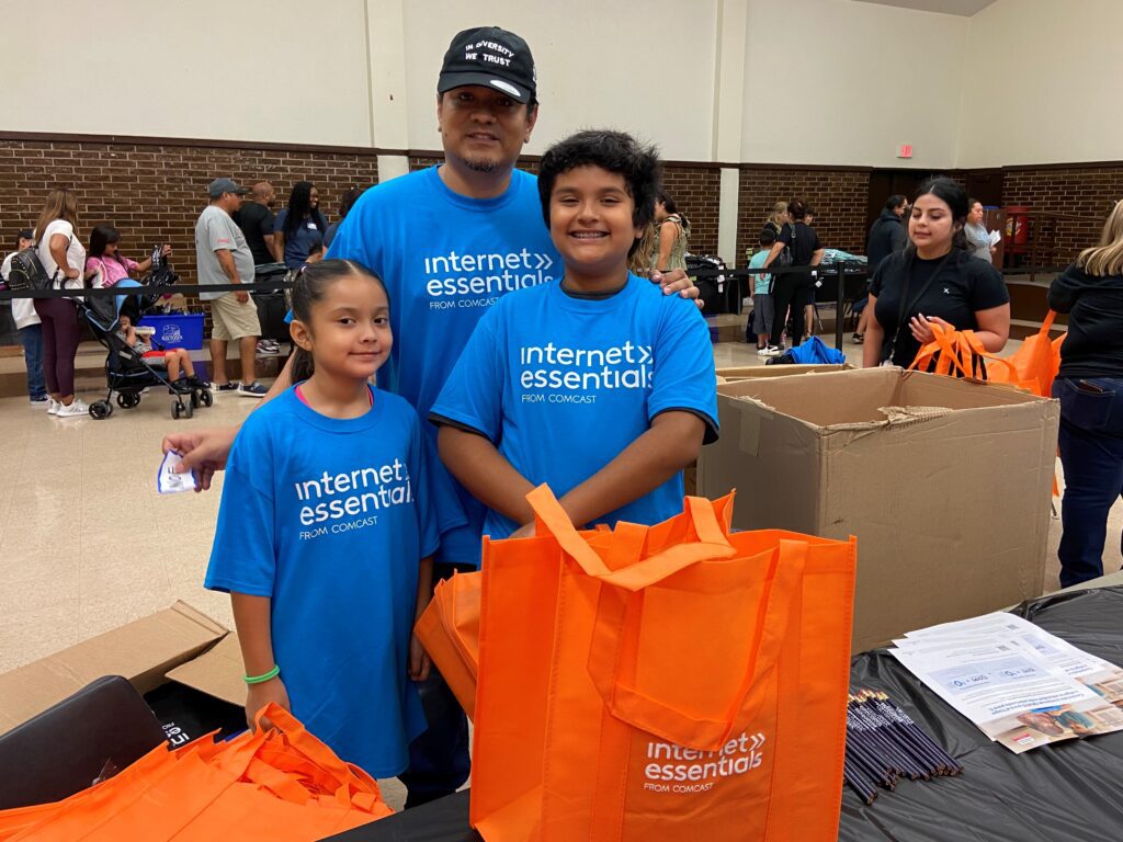 A man and two children standing near table with orange Internet Essentials bags