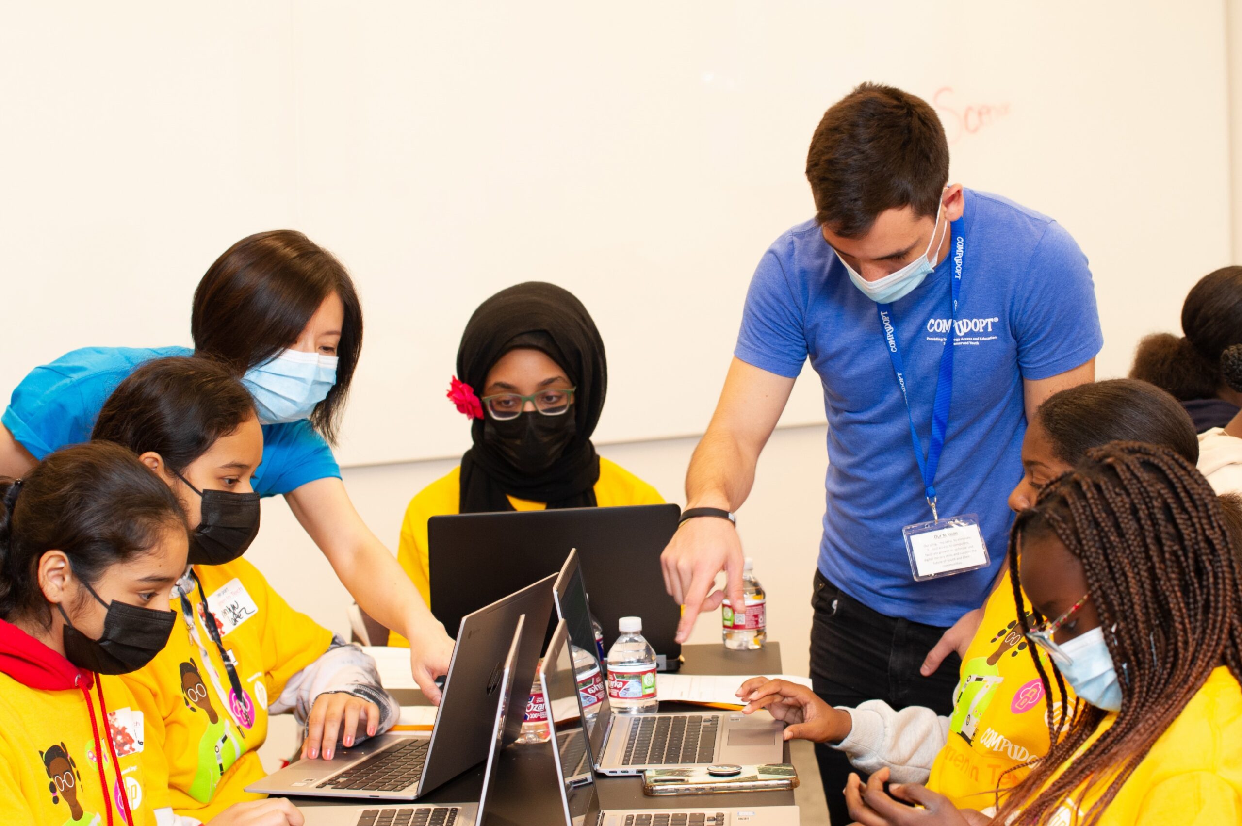 Five young girls working on laptops while two adults lean in to assist them