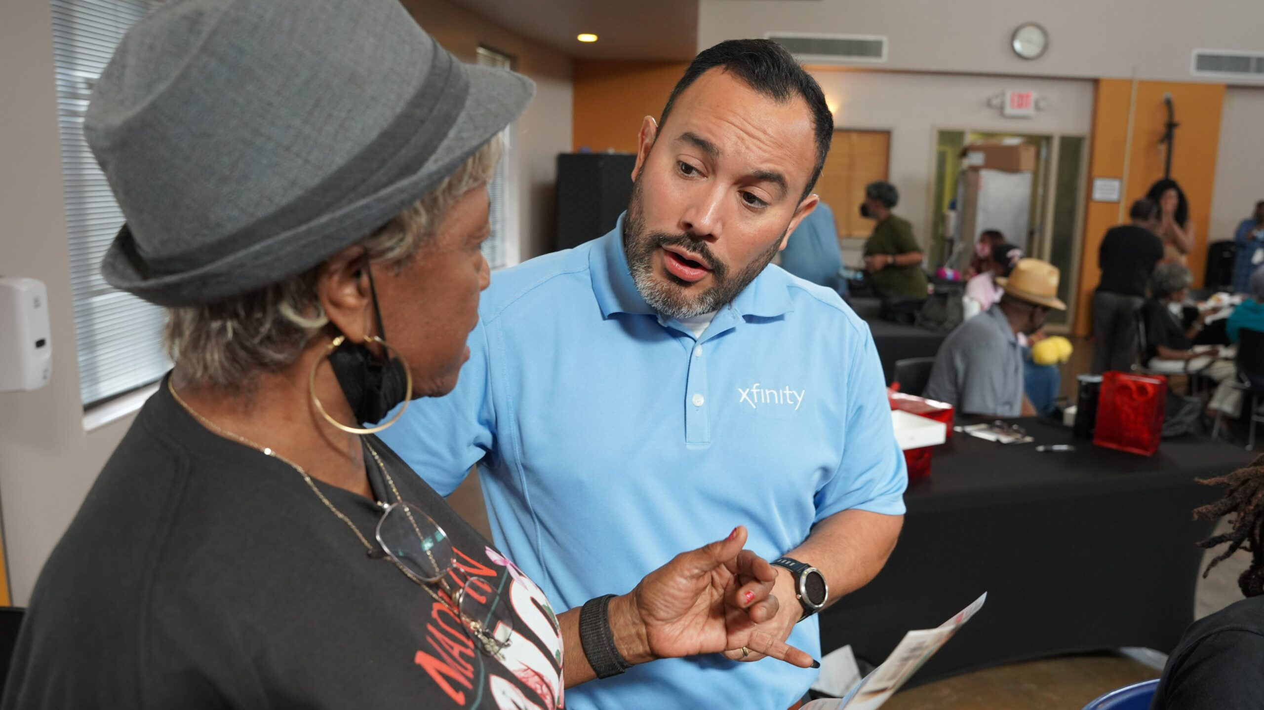 Gentleman in light blue Xfinity polo shirt speaking with older woman wearing hat.