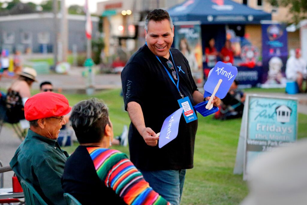 Man wearing black Xfinity shirt handing out paper xfinity fans to people seated in lawn chairs