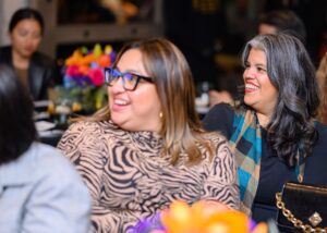 Two women seated at a table with flowers and looking to their right with smiling faces