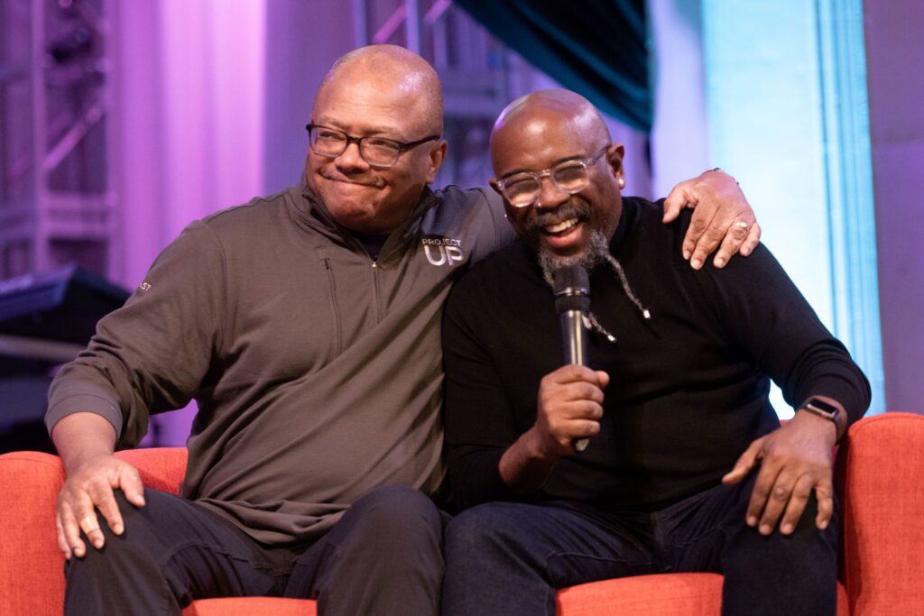 Broderick Johnson sitting with a gentleman holding a microphone.