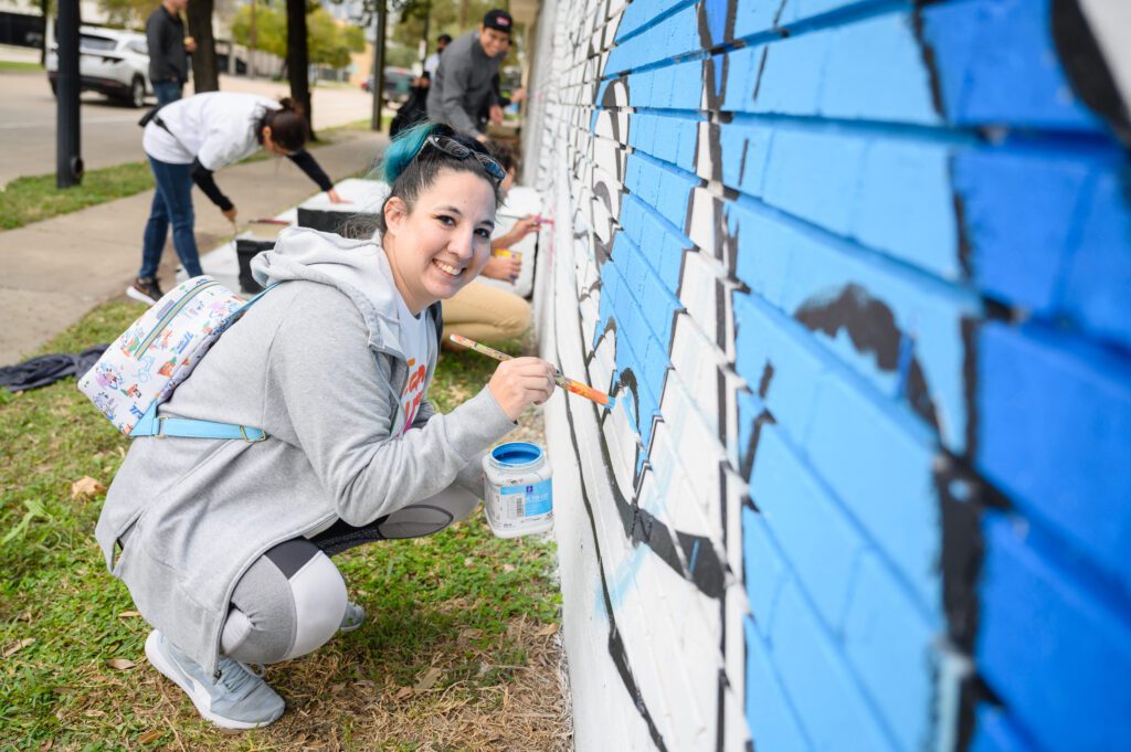 Woman wearing small backpack leaning down to paint blue on mural wall