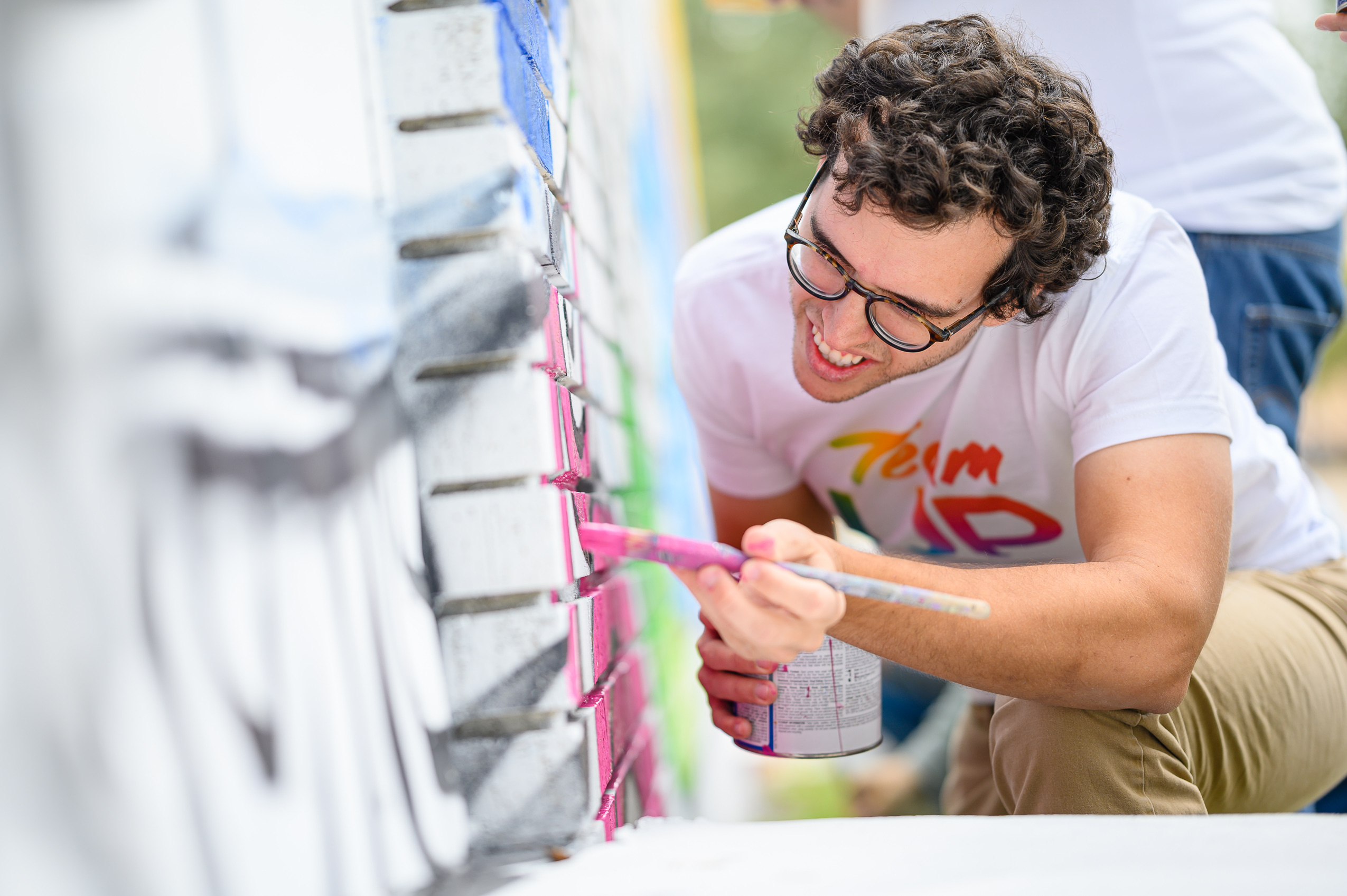 Team up volunteer wearing glasses painting on mural with red paint.