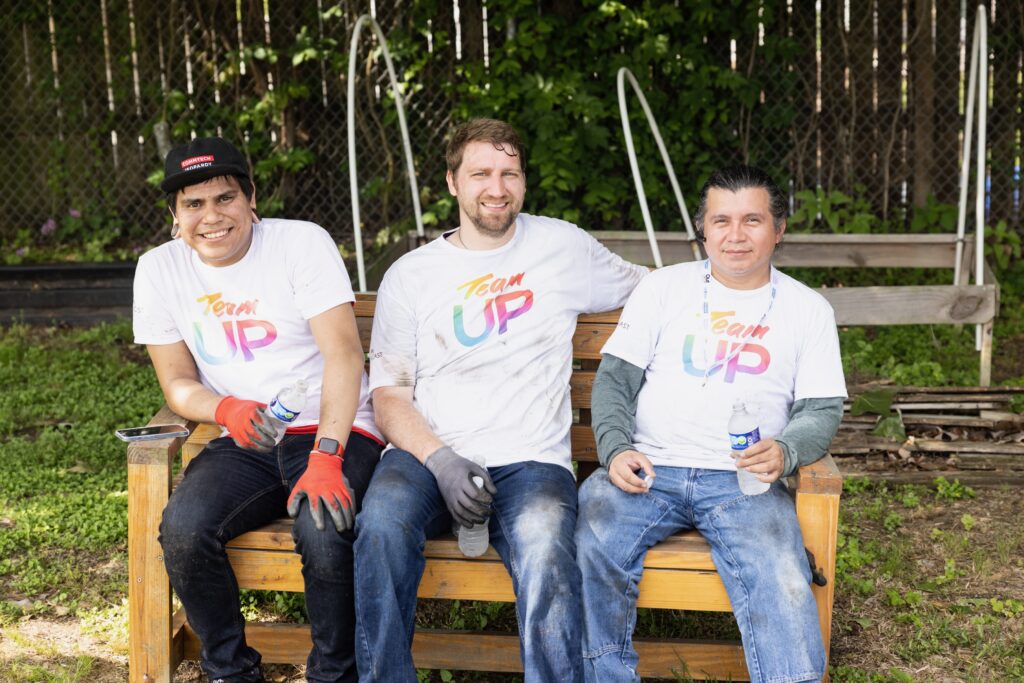 Three volunteers sitting on wooden bench with water bottles, smiling at camera