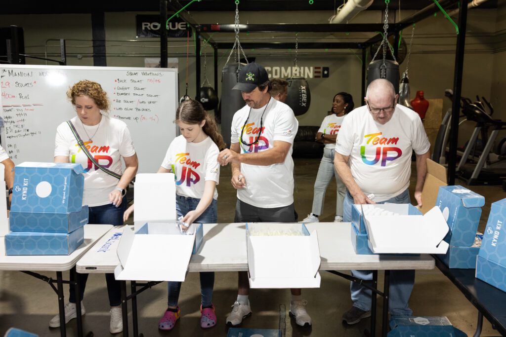 Group of volunteers standing behind a table adding items into blue boxes labels kind kits