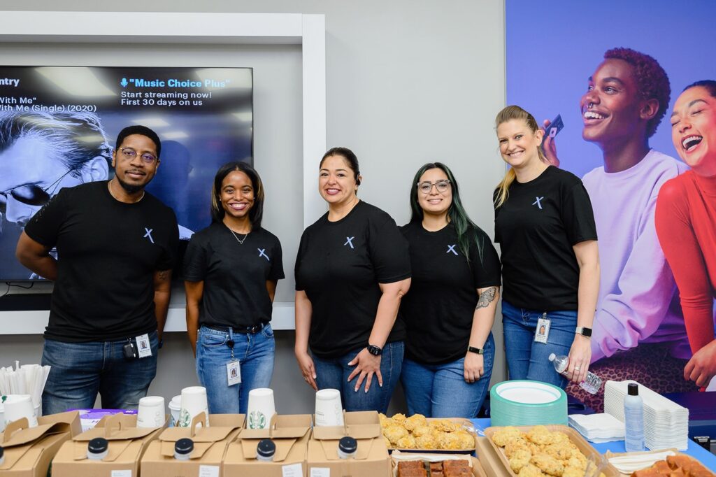 Five people wearing black Xfinity t-shirts smiling behind table filled with refreshments.