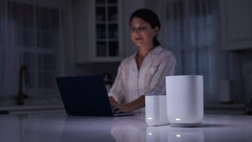 A woman sits in a dark kitchen, using her laptop on a kitchen island next to her Xfinity Storm-Ready WiFi device.