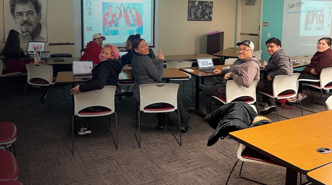 People are seen smiling sitting at desks inside a classroom.